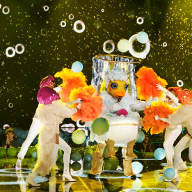 A person in a rubber duck costume in a bathtub dances onstage with four dancers in shower caps